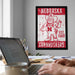 a framed poster of a cartoon character from Nebraska, with the text "NEBRASKA" and "CORNHUSKERS" prominently displayed. In the foreground, there is a laptop computer and a coffee mug on a desk, suggesting a workspace or home office setting.