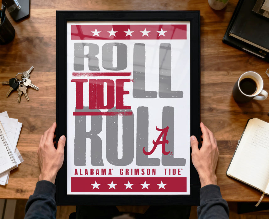 A framed poster with the text "ROLL TIDE A" is being held by a person's hands, with a wooden desk and various items in the background.