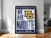 A framed poster with the text "WE ARE MARQUETTE" and a logo, placed on a wooden floor against a white wall.