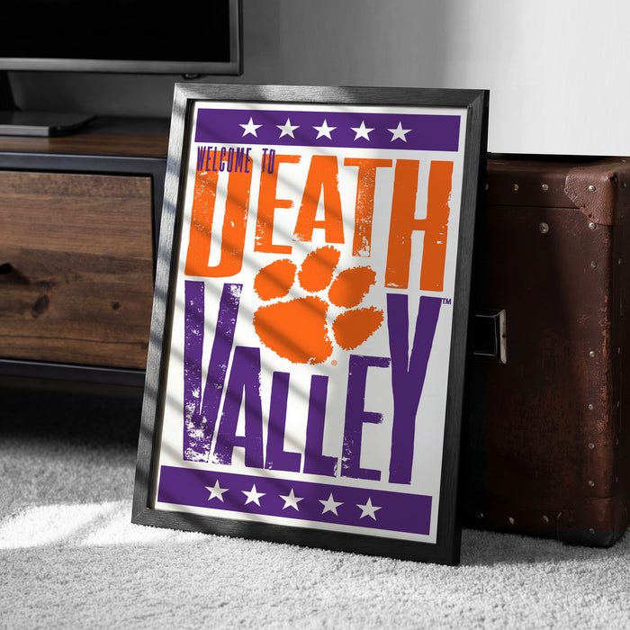 A framed poster with the text "Welcome to Death Valley" and an orange paw print graphic, placed on a carpeted floor next to a wooden chest.