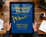 A framed poster with the text "Marquette Football" and a yellow flag, held by a person's hands.