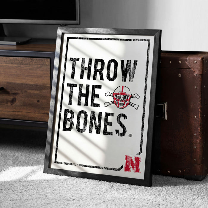 A framed poster with the text "THROW THE BONES" and a skull and crossbones symbol, placed on a carpeted floor next to a wooden chest.