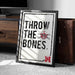 A framed poster with the text "THROW THE BONES" and a skull and crossbones symbol, placed on a carpeted floor next to a wooden chest.