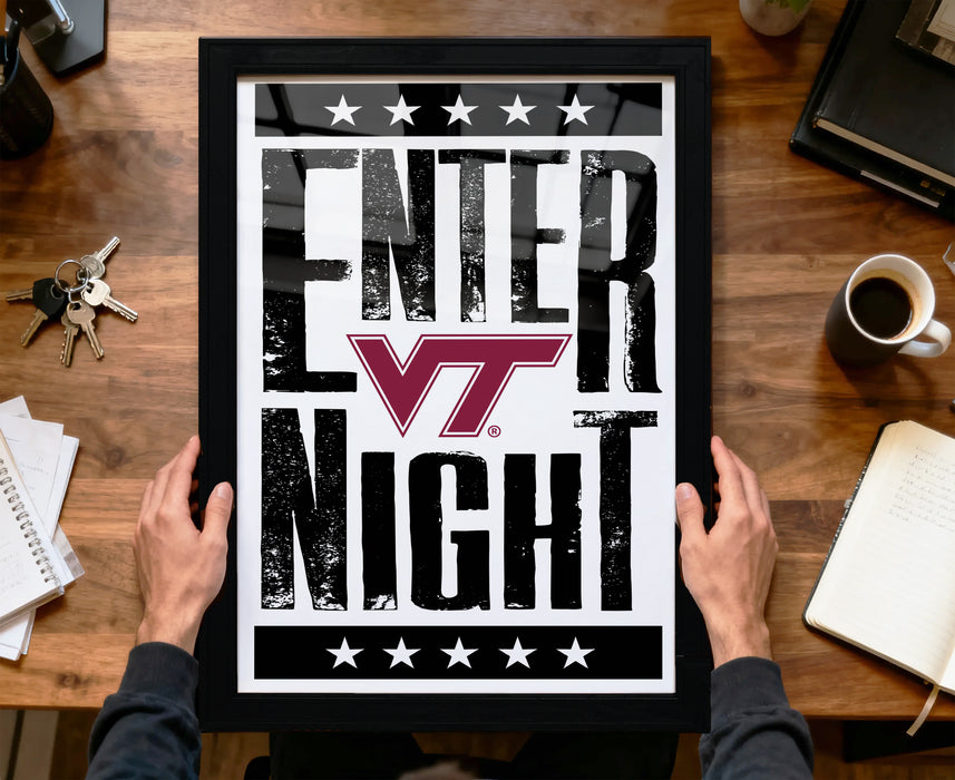 A framed poster with the text "ENTER NIGHT" is being held by a person's hands, with a wooden desk and various items such as a coffee cup, keys, and a notebook in the background.