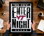 A framed poster with the text "ENTER NIGHT" is being held by a person's hands, with a wooden desk and various items such as a coffee cup, keys, and a notebook in the background.