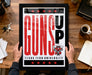 A framed poster with the text "GUNS UP" and a graphic of a character holding a gun, placed on a wooden desk.