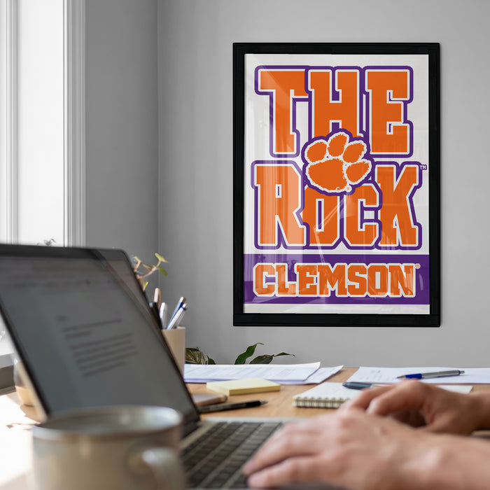 a person working on a laptop at a desk, with a framed poster of the University of Alabama's Tigers logo hanging on the wall behind them.