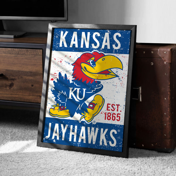 A framed poster of the Kansas Jayhawks, a Kansas football team, is displayed on a carpeted floor next to a wooden chest.