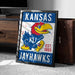 A framed poster of the Kansas Jayhawks, a Kansas football team, is displayed on a carpeted floor next to a wooden chest.