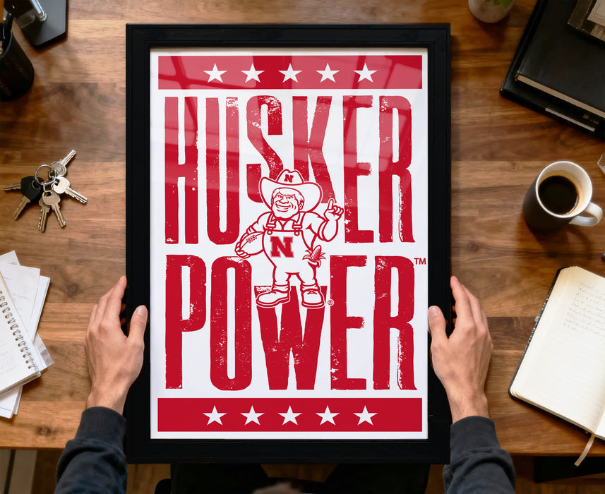 A framed poster with the text "HUSKER POWER" and a cartoon character is being held by a person's hands.