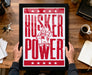 A framed poster with the text "HUSKER POWER" and a cartoon character is being held by a person's hands.