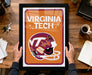 A framed poster of a Virginia Tech football helmet is being held by a person's hands, with a desk and various items in the background.