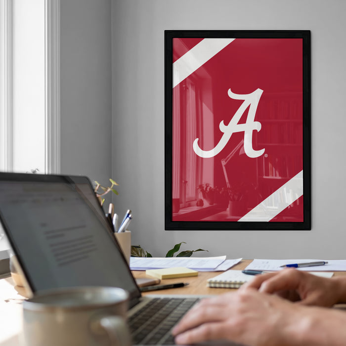 a person working on a laptop at a desk, with a framed poster of the letter "A" on the wall behind them.