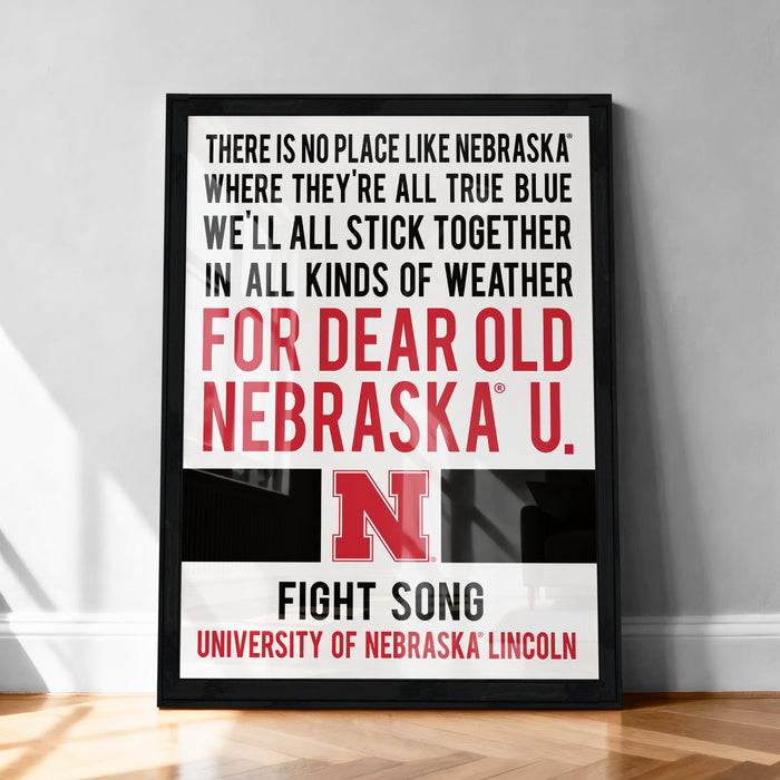 The image is a framed poster with text that reads: "THERE IS NO PLACE LIKE NEBRASKA WHERE THEY'RE ALL TRUE BLUE WE'LL ALL STICK TOGETHER IN ALL KINDS OF WEATHER FOR DEAR OLD NEBRASKA U. FIGHT SONG UNIVERSITY OF NEBRASKA LINCOLN"