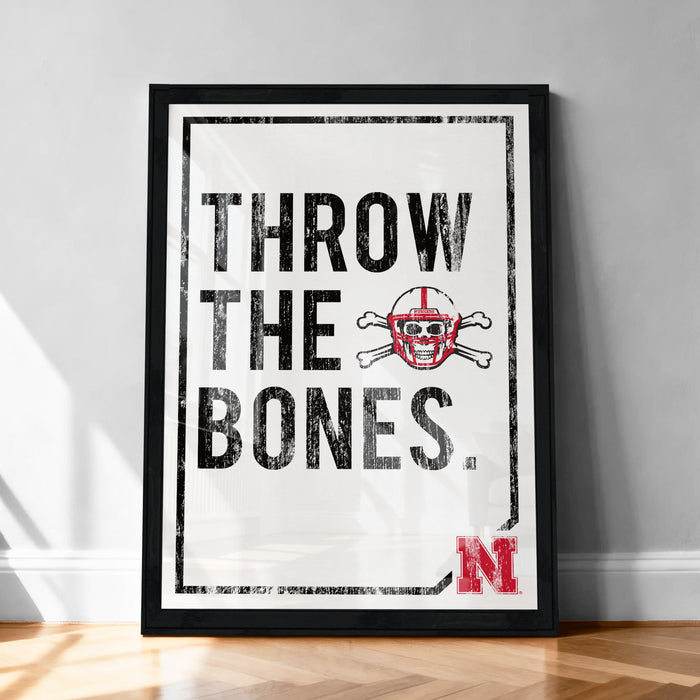 A framed poster with the text "THROW THE BONES" and a skull and crossbones symbol, placed on a wooden floor against a white wall.