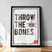 A framed poster with the text "THROW THE BONES" and a skull and crossbones symbol, placed on a wooden floor against a white wall.