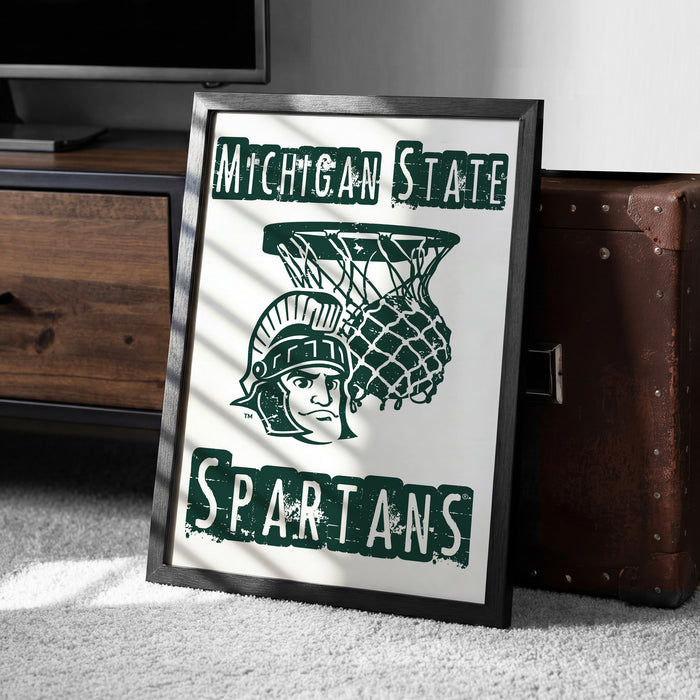 A framed poster with the Michigan State University (MSU) logo and the word "SPARTANS" printed on it, placed on a carpeted floor next to a wooden chest.