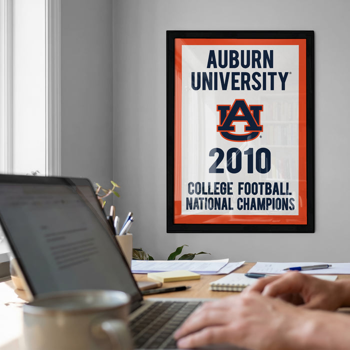 a framed poster on a wall that reads "Auburn University 2010 College Football National Champions", with a laptop and a coffee cup on a desk in the foreground.