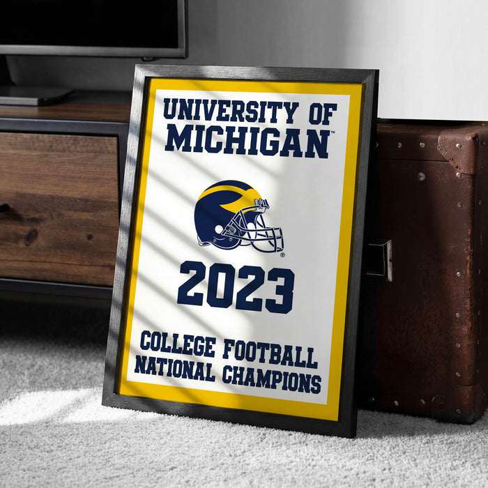 A framed poster with the text "UNIVERSITY OF MICHIGAN" and "2023 COLLEGE FOOTBALL NATIONAL CHAMPIONS" is displayed on a carpeted floor next to a wooden chest.
