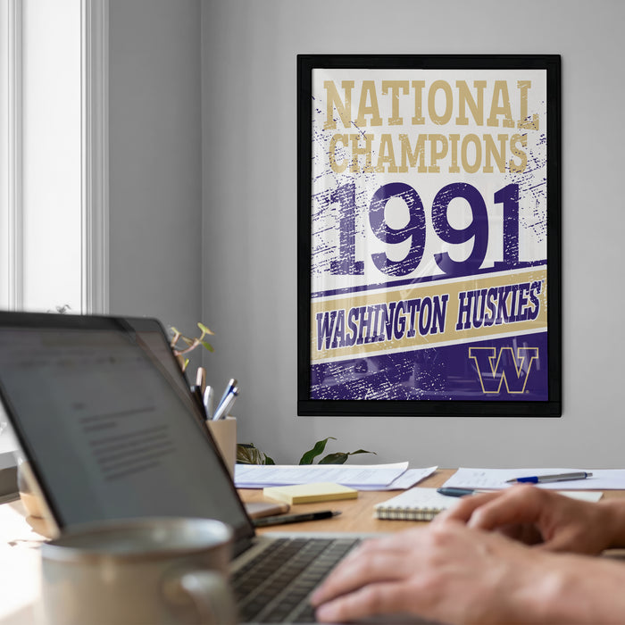 a framed poster on a wall that reads "National Champions 1991 Washington Huskies", with a person's hands visible on a laptop in the foreground.
