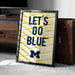 A framed poster with the text "LET'S GO BLUE™" printed on it, placed on a carpeted floor next to a wooden chest.