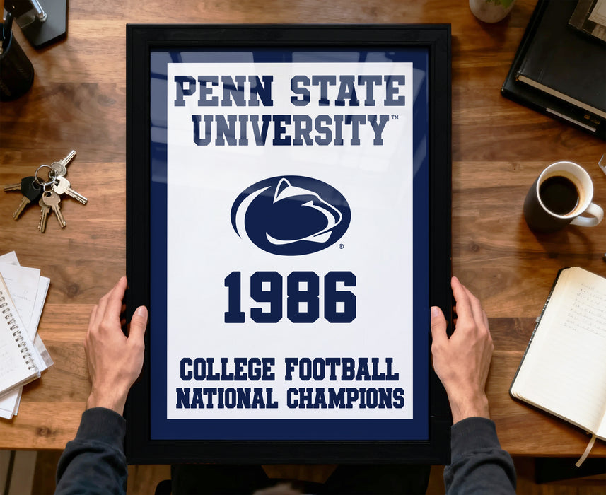A framed poster with the text "Penn State University 1986 College Football National Champions" is being held by a person's hands, with a keyboard, coffee cup, and notebook visible in the background.