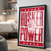 A framed poster with the text "HUSKER POWER" and an illustration of a character wearing a cowboy hat and holding a sign.