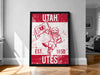 A framed poster of a red and white design featuring a bird-like character holding a flag, with text that reads "UTAH EST. 1850 Utes".