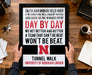 A framed motivational quote is being held by a person's hands, with a wooden desk and various items in the background.