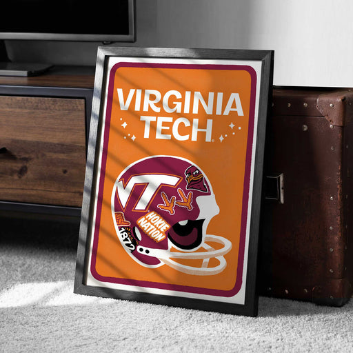 A framed poster of a Virginia Tech football helmet is displayed on a carpeted floor next to a wooden chest.