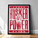 A framed poster with the text "HUSKER POWER" and an illustration of a character wearing a cowboy hat and holding a sign.