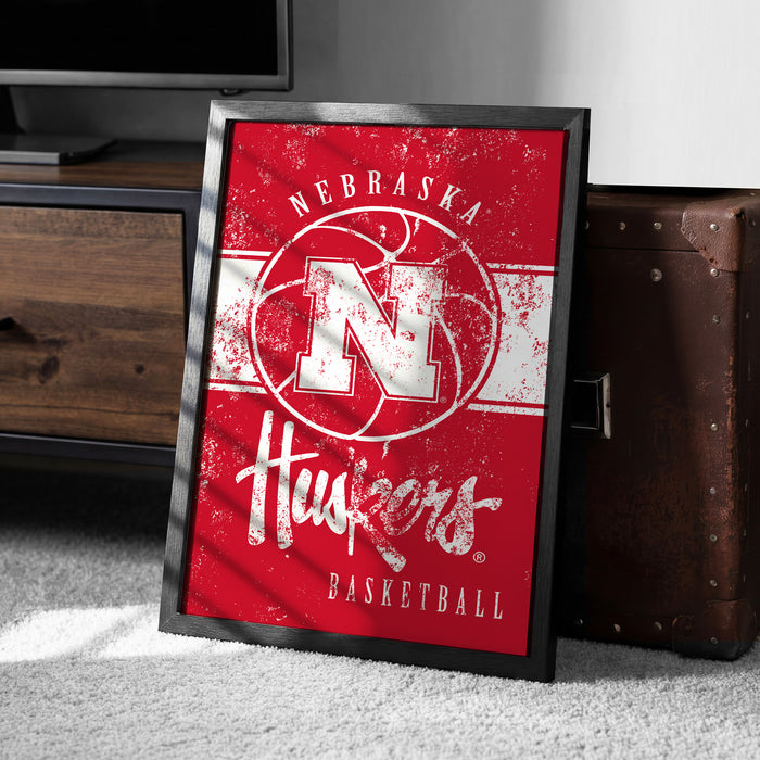 A framed poster of the Nebraska Huskies basketball team, featuring the team's logo and text, is displayed on a carpeted floor next to a wooden chest.