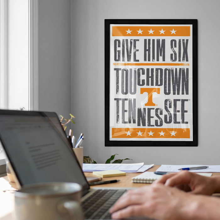 a person working on a laptop at a desk, with a framed poster on the wall behind them that reads "GIVE HIM SIX TOUCHDOWN TENNESSEE".