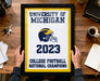 A framed poster titled "UNIVERSITY OF MICHIGAN" with a football helmet and the year "2023" displayed, along with the text "COLLEGE FOOTBALL NATIONAL CHAMPIONS".