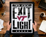 A framed poster with the text "EXIT LIGHT" is being held by a person's hands, with a wooden desk and various office items in the background.