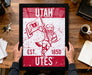A framed poster of a red and white design featuring a bird character holding a flag, with text that reads "UTAH EST. 1850 Utes".
