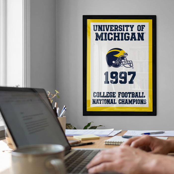 a framed poster of the University of Michigan football team celebrating their 1997 College Football National Champions, with a laptop and a person's hands visible in the foreground.