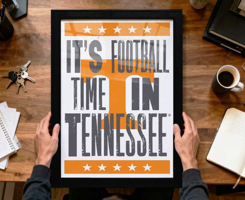 A framed poster with the text "IT'S FOOTBALL TIME IN TENNESSEE" is being held by a person's hands, with a wooden desk and various items such as a cup, keys, and a notebook in the background.