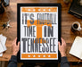 A framed poster with the text "IT'S FOOTBALL TIME IN TENNESSEE" is being held by a person's hands, with a wooden desk and various items such as a cup, keys, and a notebook in the background.