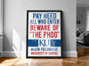 A framed poster with the text "PAY HEED ALL WHO ENTER BEWARE OF THE PHOG" and the university's logo, placed on a wooden floor against a white wall.