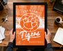 A framed poster of the Clemson Tigers basketball team is being held by a person's hands, with a desk and various items such as a coffee cup, keys, and a notebook visible in the background.