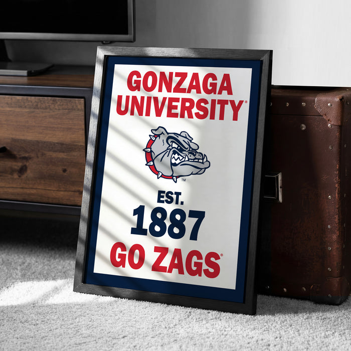 A framed poster with the text "GONZAGA UNIVERSITY EST. 1887 GO ZAGS" and a bulldog logo, placed on a carpeted floor next to a wooden chest.