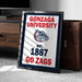 A framed poster with the text "GONZAGA UNIVERSITY EST. 1887 GO ZAGS" and a bulldog logo, placed on a carpeted floor next to a wooden chest.
