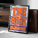 A framed poster with the text "THE ROCK CLIMB" and "CLAMSON" printed on it, along with an orange paw print symbol, is leaning against a wooden cabinet.